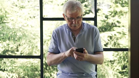Mature man using smartphone standing by window at home
