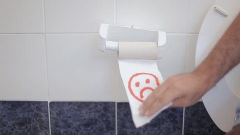 A man's hand picks the last piece of paper from a toilet roll and discovers a sad emoticon drawn in red ink. Medium shot.
