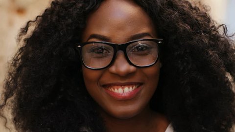 Close up portrait of beatiful curly hair african woman in the glasses smiling on the camera on the street background.