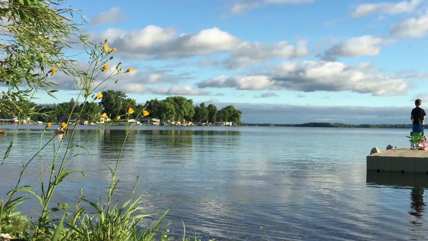 People canoe by on glassy water while little boy is fishing from dock; shot in late afternoon in summer on Minnesota lake.