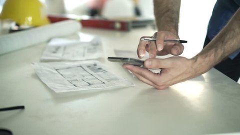 Man working with smartphone and blueprints by table at his new home

