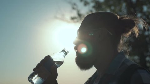 Slow motion shot of side view of stylish man with ponytail drinking water from bottle on background of sunlight and nature.
