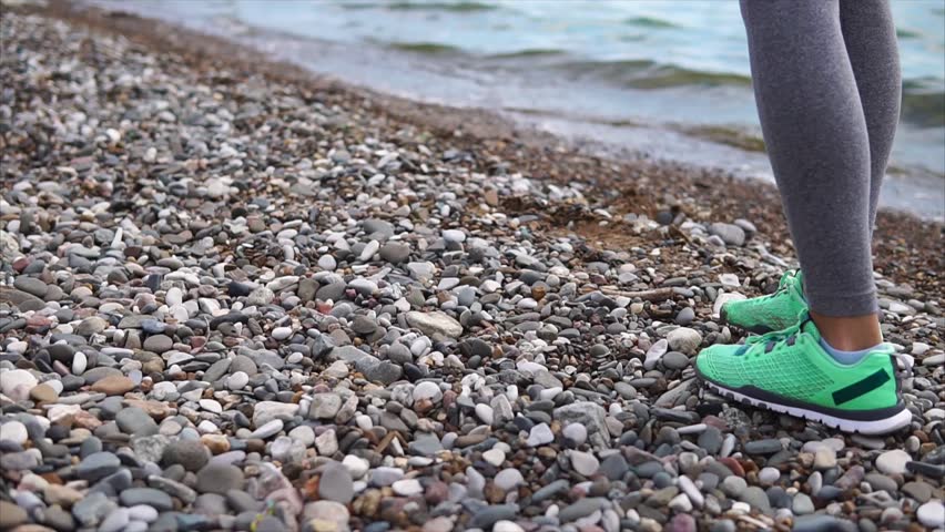close up shot of the feet of a young woman who dressed in sports sneakers, she runs along the stony bank near the river or the sea, the lady is engaged in sports in the summer