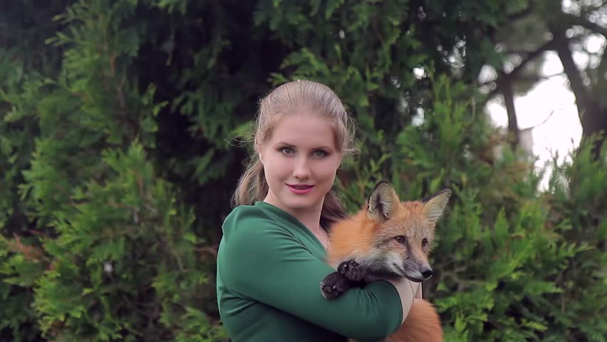 Young woman holding fox and posing front of camera outdoors. She is keeping her beloved red pet and looks with smile, standing in street against background of green trees. Photoshoot with fox is