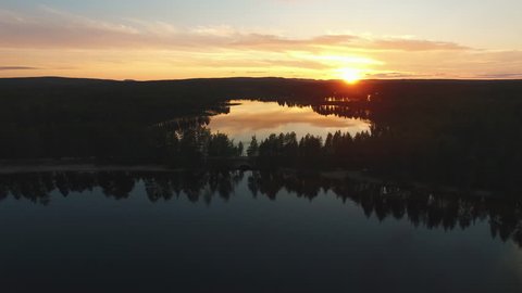 Aerial shot of lake and trees at sunset. Flying over calm lake and road in forest at dusk, reflections in the water