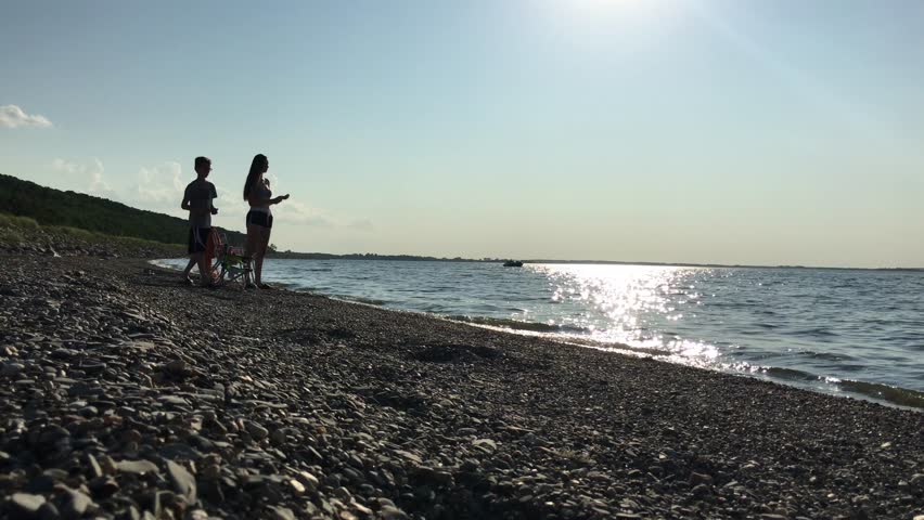 Kids skipping rocks and mostly silhouetted by the beaming background sun that is partially in the shot and reflecting uniquely off of the water; shot on a rocky beach on Devils Lake in North Dakota.