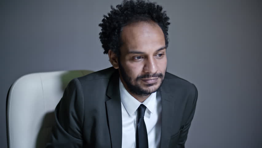 African businessman in formal suit and tie sitting in dark office and speaking with somebody