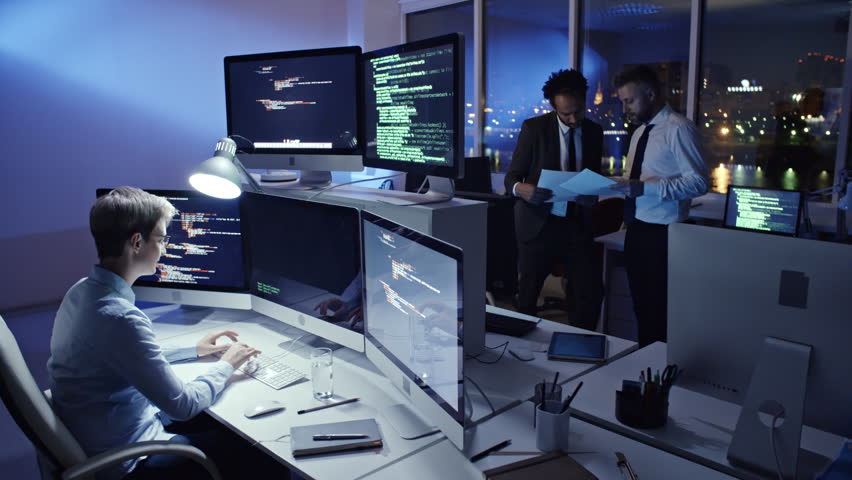 Female programmer writing computer code on computer workstation in the office at night while her male colleagues discussing documents Royalty-Free Stock Footage #29974138