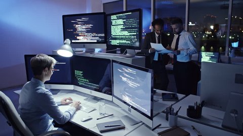 Female programmer writing computer code on computer workstation in the office at night while her male colleagues discussing documents