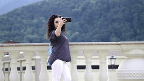 Smart, beautiful brunette corrects curly hair and makes selfie on a mobile phone against the background of outstanding mountains and beautiful sunset.