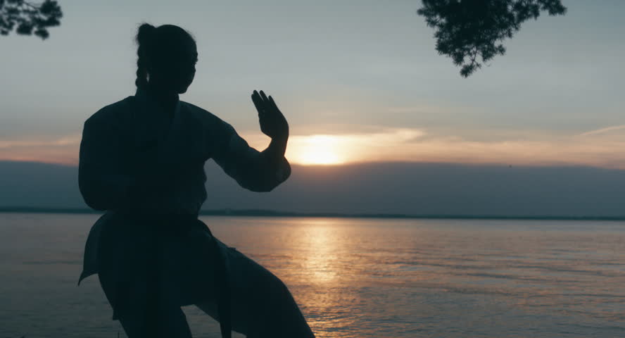 MED Caucasian professional female athlete wearing kimono practicing karate near large lake, sunset shot. 4K UHD, 60 FPS SLO MO
