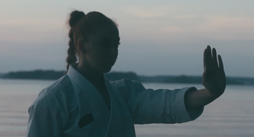 CU Caucasian professional female athlete wearing kimono practicing karate near large lake, sunset shot. 4K UHD, 60 FPS SLO MO