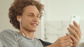 Handsome cheerful man with curly hair and headphones lying on sofa and chatting on smartphone
