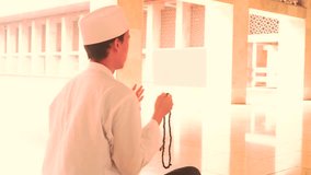 Video footage of a young muslim man praying with beads while sitting in the Istiqlal mosque and wearing islamic clothes
