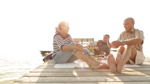 dolly shot four seniors enjoying picnic on jetty at lake on late summer day
