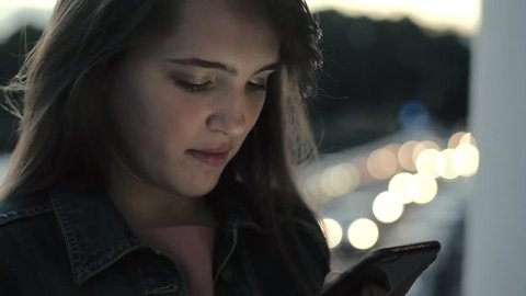Smiling young woman typing message on mobile phone on background evening city. Close up girl chatting and looking cell phone on background evening lights city. Portrait young woman using phone