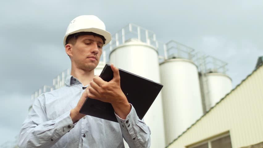 An engineer in white helmet with a tablet in his hands the backdrop of a factory