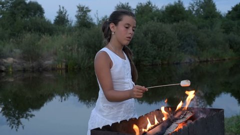 A pretty little girl fries marshmallows on the fire near the pond. camping, travel, tourism, hike and people concept - happy family roasting marshmallow over campfire. Toasting Marshmallows