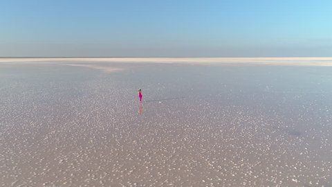Lonely young caucasian woman in red dress with long hairs is walking on surface of huge salt lake. Drone is flying around. Aerial view