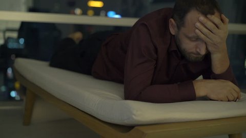 Sad, unhappy man lying on lounger at home at night
