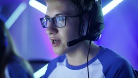 Professional Boy Gamer Plays in Video Game on a eSports Tournament/ Internet Cafe. He Wears Glasses and Headphones and Speaks into Microphone. Other Girls and Boys Players Playing in Background. 