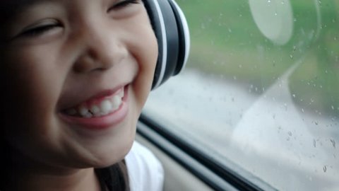 Asian pretty girl listening to music with headphones moving in car and looking away in rainy day