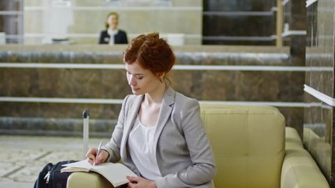 Tracking of young businesswoman sitting in armchair and writing in organizer while businessman walking through hotel lobby in slow motion