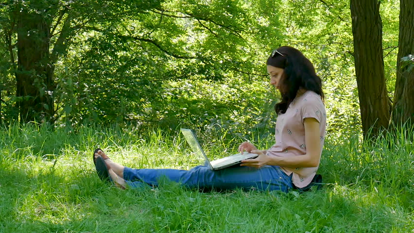 Outdoors Portrait of Young Brunette Woman with White Laptop Sitting on Green Grass in the Park and Working with Pleasure. Mobility Concept.