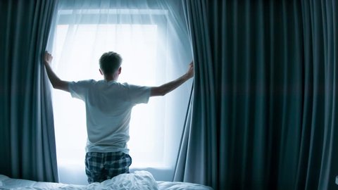 Middle aged Caucasian man opening curtains looking through window in a hotel room beginning of a new day