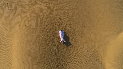 Young woman in white dress lies on golden sand in desert. Aerial vertical top shot. Drone is spinning around and flying upward