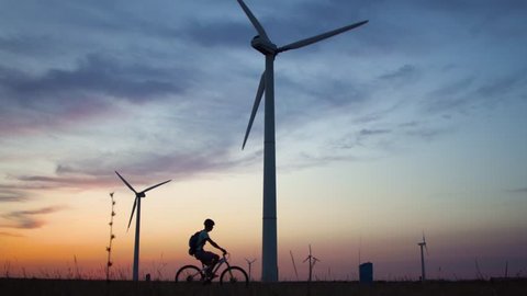 A young man on a bicycle rides past a wind power plant at sunset