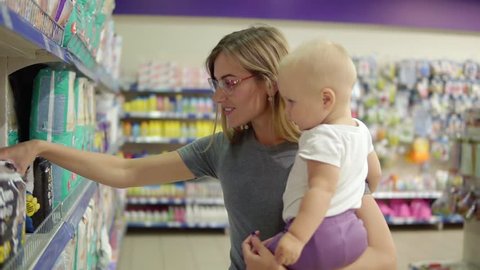 Young attractive mother in glasses holding her child in her arms while choosing diapers on the shelves in the supermarket. Thoughtful mom carefully choosing best products for her child