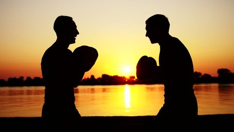 Two dark male figures, at sunrise, against the light, boxing, fighting in sparring, training in a pair of techniques of strikes. On sandy beach, in cargo port, near water, in summer