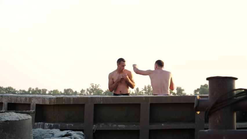 Young athletic men with bare, naked torsos, box, practice the technique of strikes, capture, fight, on a sandy beach, in a cargo port, near large cargo cranes, at sunrise, in summer