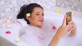 Video footage of a young woman relaxing on bathtub while using a cellphone and smiling happy