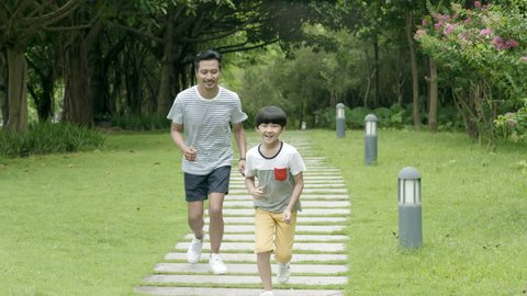 asian father running behind his son in park in summer in slow motion