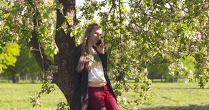 slow motion slide shot of teenage girl standing under blossoming pink apple tree talking on the phone