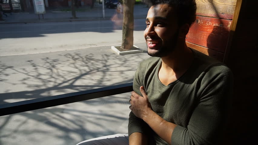 Boy sitting on sofa and puts hand on hand. Attractive male has dimples, beard, full lips and black hair. Handsome boy dressed in khaki pullover. Concept of new modern places for resting fast customer