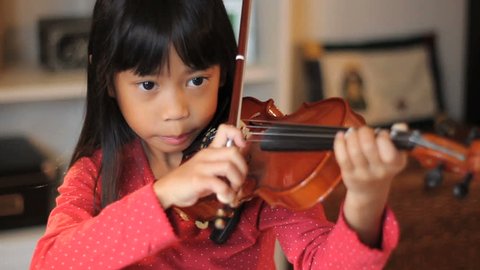 A pretty 6 year-old Asian girl diligently practices her violin in the living room.