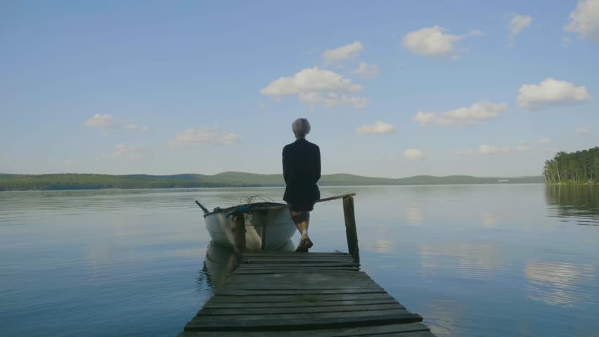 Back view of a woman sitting on a tiny wooden pier by the pond. Young beautiful girl sitting on jetty river on background. Back view