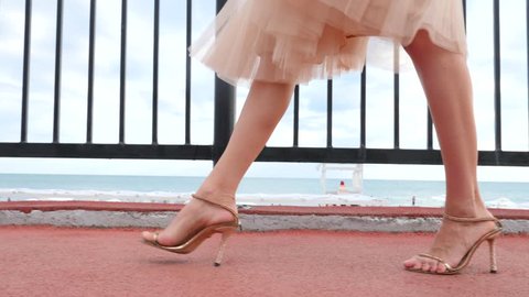 An elegant woman in a hat and a white air dress strolls along the waterfront. Legs close-up. 4k, slow-motion