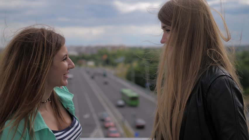Close-up of two cute and laughing young women say goodbye to each other. They embrace, kiss each other and wave their hands. Slow motion, the hair develops in the wind. On the street.