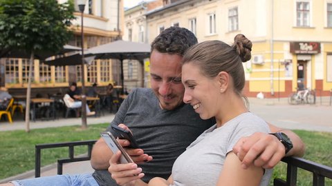 Happy young couple sitting on a bench and using smart phone. Woman and man watching video, photos