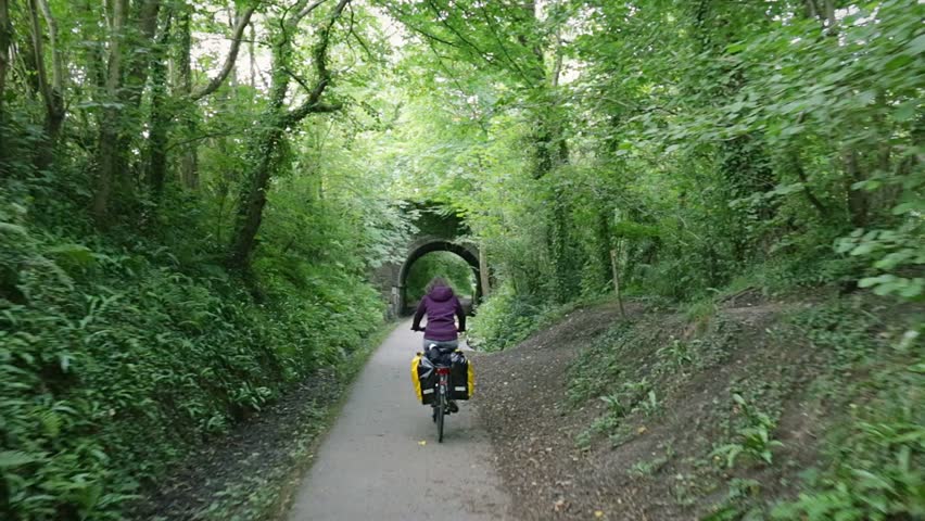 Woman on a bike drive bicycle under stone bridge with a lush green forest around. POV behind the cyclist