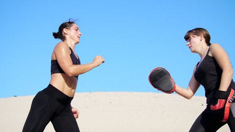 Two athletic, young women in black fitness suits are engaged in a pair, work out kicks, train to fight, on deserted beach, against a blue sky, in summer, under a hot sun. Slow motion