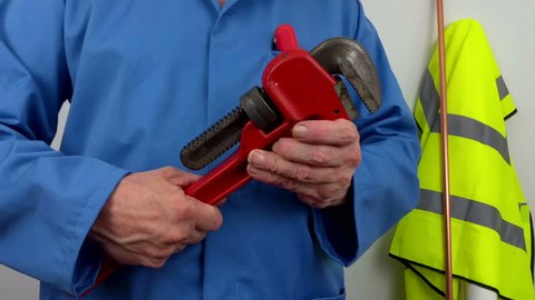 Plumber in blue overalls opening an adjustable wrench standing next to a hi vis vest hanging up
