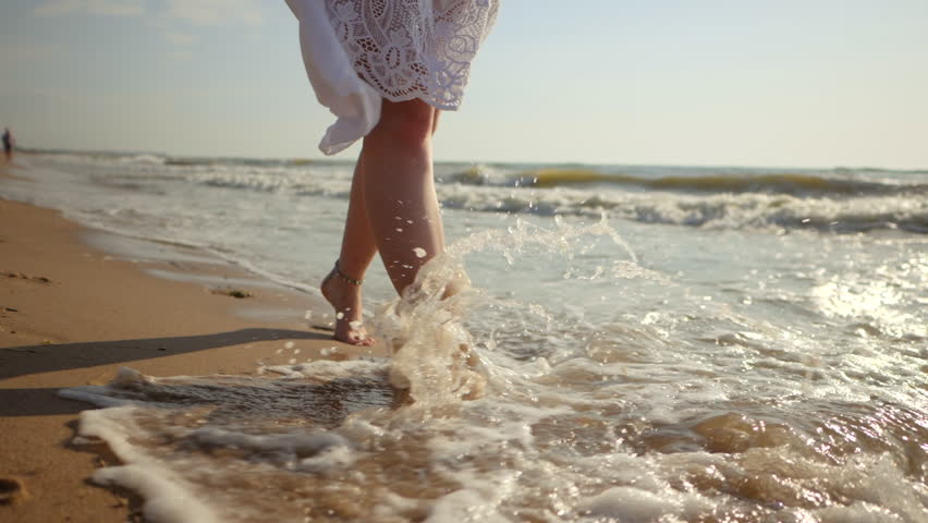 Legs of caucasian girl wearing white long dress and silver bracelets walking barefoot sand on sea beach, sun, lens, flare. Slow motion. Woman walks towards to camera | Shutterstock HD Video #30222091