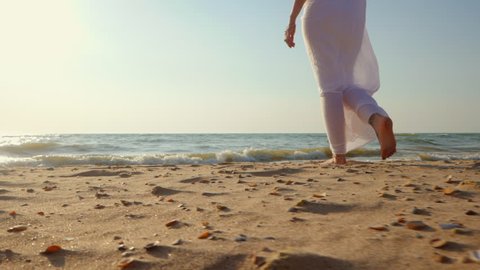Legs of caucasian girl wearing white long dress and silver bracelets walking barefoot sand on sea beach, sun, lens, flare. Slow motion. Woman walks away camera