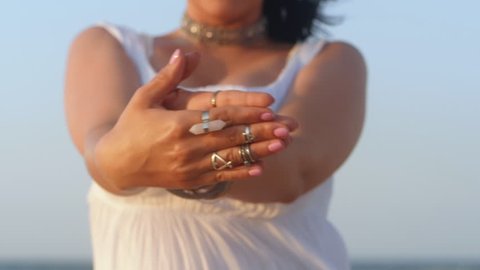 Beautiful young gypsy woman dancing hands with boho jewelry. Girl in white long dress showing bohemian accessories near sea on the beach. Slow motion. Femininity concept.