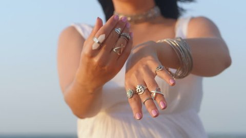 Beautiful young gypsy woman dancing hands with boho jewelry. Girl in white long dress showing bohemian accessories near sea on the beach. Slow motion. Femininity concept.